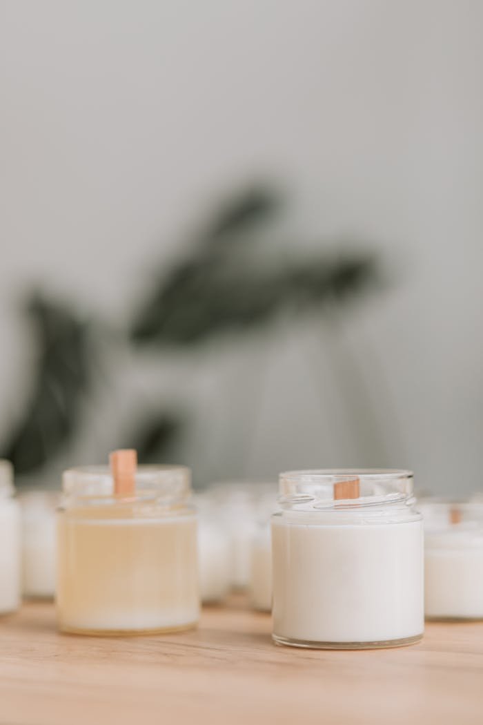 Close-up of handmade candles with wooden wicks in glass jars on a wooden surface.